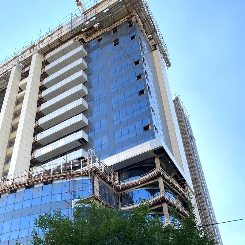 Glass tower under construction seen from below, white fins rising above a curved glass podium, trees in the foreground.