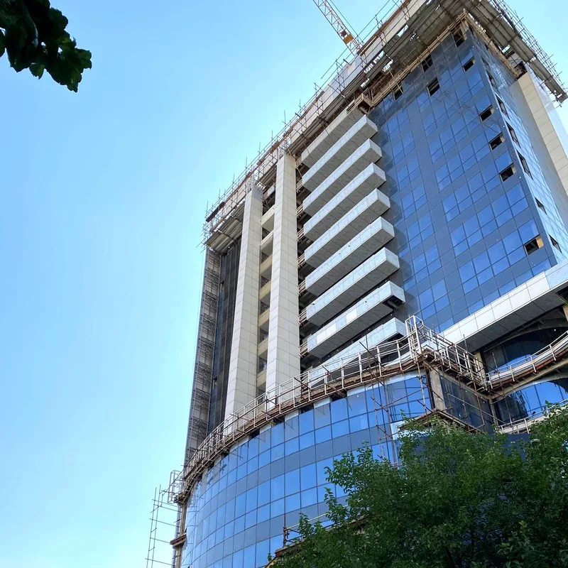 A tower crane above a glass tower under construction, its curved glass podium wrapped in scaffolding.
