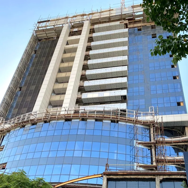 Curved blue glass podium of a tower under construction, with scaffolding on the facade above it.